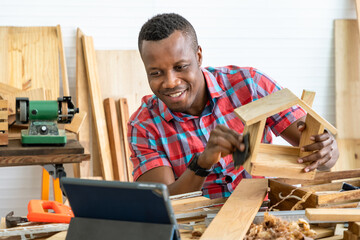 African black male carpenter man learning from internet with tablet, and sanding wooden house model toy with sandpaper in carpentry workshop. Concept hobby at home.