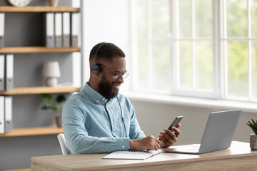 Satisfied attractive young black guy with beard in glasses and headphones chatting on phone at workplace with pc