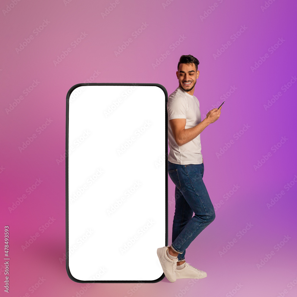 Poster Happy young Arab man using mobile device while leaning on big smartphone with empty screen in neon light. Mockup