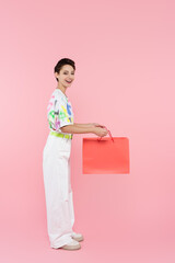 full length view of happy woman in white trousers holding shopping bag on pink