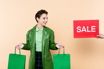 man holding red card with sale lettering near trendy woman with green shopping bags on beige