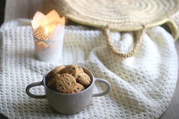 Bowl of chocolate chip cookies, soft knitted basket, wicker basket and candle holder with lit candle. Hygge at home. Selective focus.