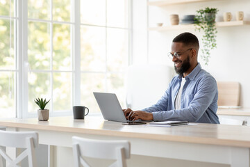 Glad millennial african american man with beard in glasses chatting remotely, work on computer alone