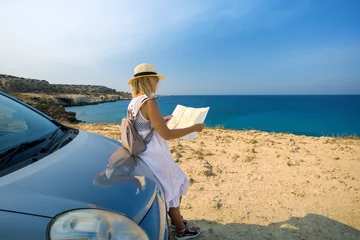 Zelfklevend Fotobehang A beautiful and stylish woman is traveling by car. Mature female traveler travel  by car, look at the road map against the sea, Cyprus. © Sergey