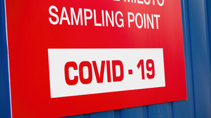 Detail of red label saying COVID-19 sampling point. Blue metallic container for medical testing for infectious virus causing pandemic outbreak. Sign with coronavirus.