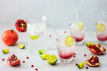 Refreshing pomegranate drink in glasses with pomegranate seeds and ice, lime and glass straws, jug of cold water with citrus, cut pomegranates with seeds
