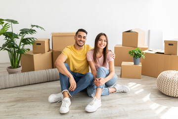 Full length portrait of loving multinational couple sitting on floor among cardboard boxes, hugging...