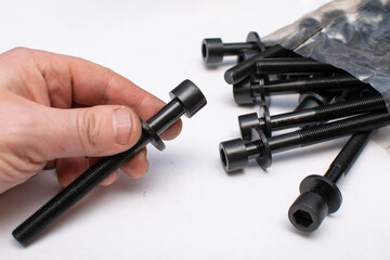 a black bolt from a car repair kit in a man hand on a white light table against a background of packaging with many identical new bolts from the cylinder head of the car engine