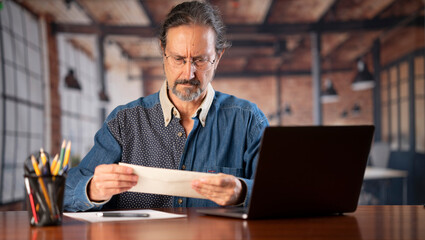 Fototapeta premium Angry stressed businessman opening envelope reading bad news in mail letter. Mad man feels frustrated about high bills, dismissal notice, bank debt, tax invoice or mistake sits at home office desk.