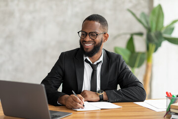 Obraz premium African American man in glasses using laptop writing in notebook
