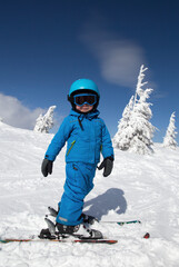 Fototapeta na wymiar toddler boy in helmet, goggles, blue overalls stands on skis on snowy mountain slope on sunny cold winter day. Children's skiing lesson at ski school. Winter active entertainment , sports education