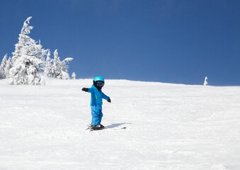 toddler boy spreading his arms stands on skis on a snow-covered mountain slope on a sunny cold winter day. Children's skiing lesson at the ski school. Winter active entertainment sports education.