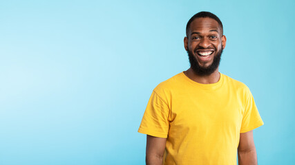 Positive young black man in casual wear smiling and looking at camera on blue background, panorama with empty space