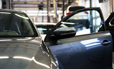 Close-up shows the front of a car with an open door which is in a car service. In the background there is an interior of a car service and several cars. The background is blurred.