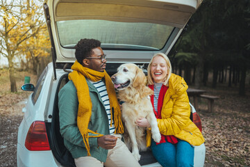 Photo of a couple on a road trip with their dog. Photo of a young couple in love and their dog sitting the trunk of a car on a beautiful autumn day; taking a short break during their road trip.