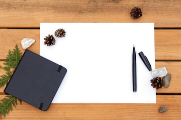 Top view of blank paper page on wood background with different objects - black, open marker, green branch, black notebook, conifer cones, stone. Flat lay style with empty space for text.