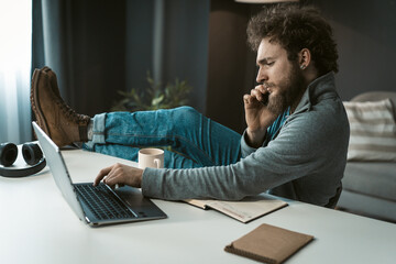 Brutal Businessman has a Serious Telephone Conversation with Suppliers and is Typing on a Computer. Feet on the Table. Work Desk with Laptop, Diary and White Cup. Male Profile. Copy Space. 