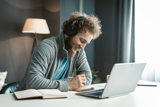 Young Student has an Online Video Lecture Uses a Laptop and Headphones, Takes Notes in a Notebook. Curly-Haired Guy sits in Earphones at the Table in his Office. Close-up