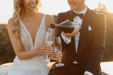 young couple, bride and groom, spend romantic evening after wedding day on rooftop against backdrop of city, man carefully pours champagne or wine into glasses