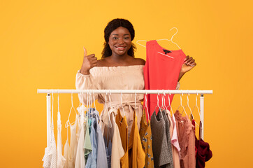 Attractive young black woman holding dress near clothing rack, showing thumb up gesture, recommending new trendy look