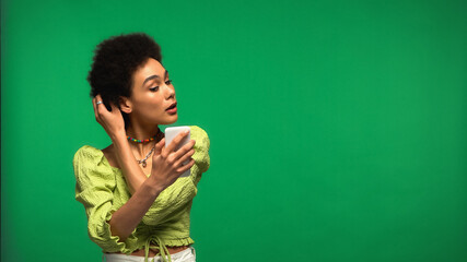 african american woman in blouse looking at smartphone and adjusting hair isolated on green