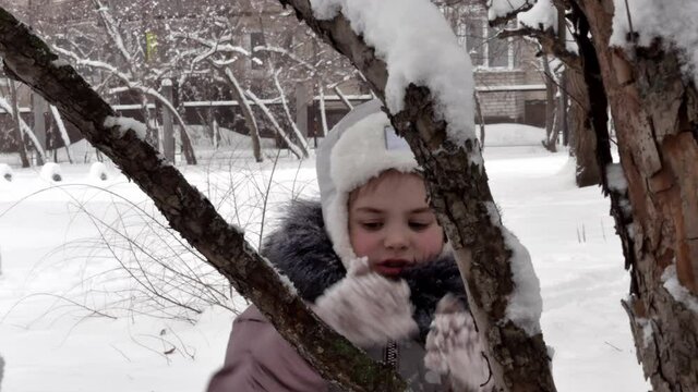 A beautiful happy little girl in winter in warm clothes, a down jacket and a hat with earflaps is having fun among the trees, hugging a branch. Street walk child happy childhood.