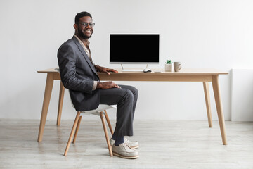 Handsome young black businessman in formal suit sitting near office table with empty computer monitor, offering mockup