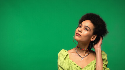 dreamy african american woman in hoop earrings adjusting curly hair isolated on green