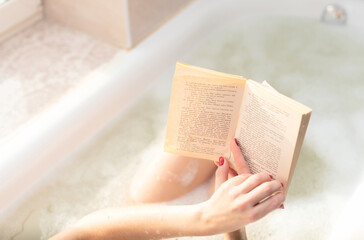 Young woman takes a bath with lush foam near the window reading the book close up. Personal hygiene, healthcare, relax at home concept. Selective focus