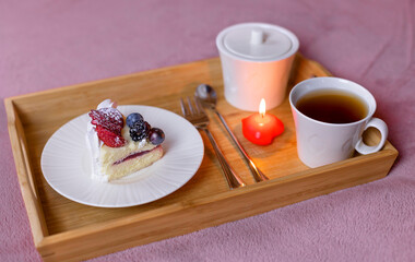 A delicious cake on a white plate and a wooden tray with a mug of tea and a candle in the shape of a heart, standing on a soft blanket on the bed.