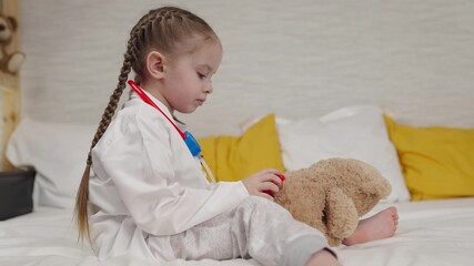 little kid plays with teddy bear sitting on bed in hospital, childhood dream of becoming doctor, kid veterinarian examines toy doll with stethoscope, pretend heartbeat, little daughter treats friend