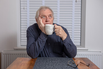 Bored senior man sitting at dining table drinking morning coffee 