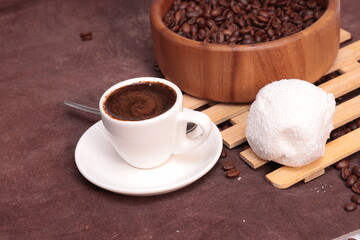 White cup with brewed coffee and coffee beans in a wooden plate. Freshly brewed espresso in a white cup surrounded by roasted coffee beans on a rustic wooden table.