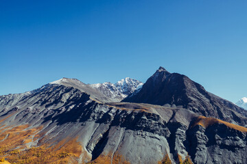 Scenic alpine landscape with sharp rocky pinnacle and snow-covered mountain in sunlight in autumn. Motley autumn scenery with gray black orange mountain with peaked top in sunshine under blue sky.