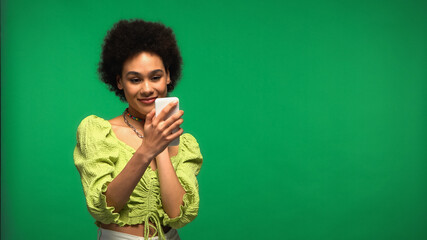 Fototapeta premium pleased african american woman in blouse looking at smartphone isolated on green