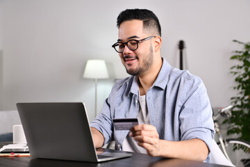 Feeling happy and positive. Young Asian man using laptop computer holding credit card for online shopping and payment at home.