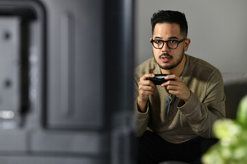 Feeling excited and engaging. Young asian man playing video game with mouth slightly opened in...