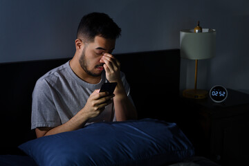 Young Asian man having sore and tired eyes when using smartphone while lying in bed at night. Young...