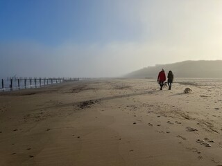 Beautiful landscape with female couple walking with dog on misty environment on sandy beach at Happisburgh in Norfolk coast in East Anglia uk with blue skies after fogbow on bright morning in December © Carmina