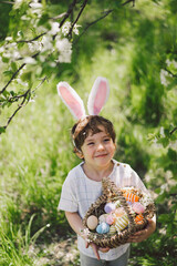 Easter egg hunt in spring garden. Funny boy with eggs basket and bunny ears on Easter egg hunt in sunny spring garden. Children celebrating Easter. Happy easter card