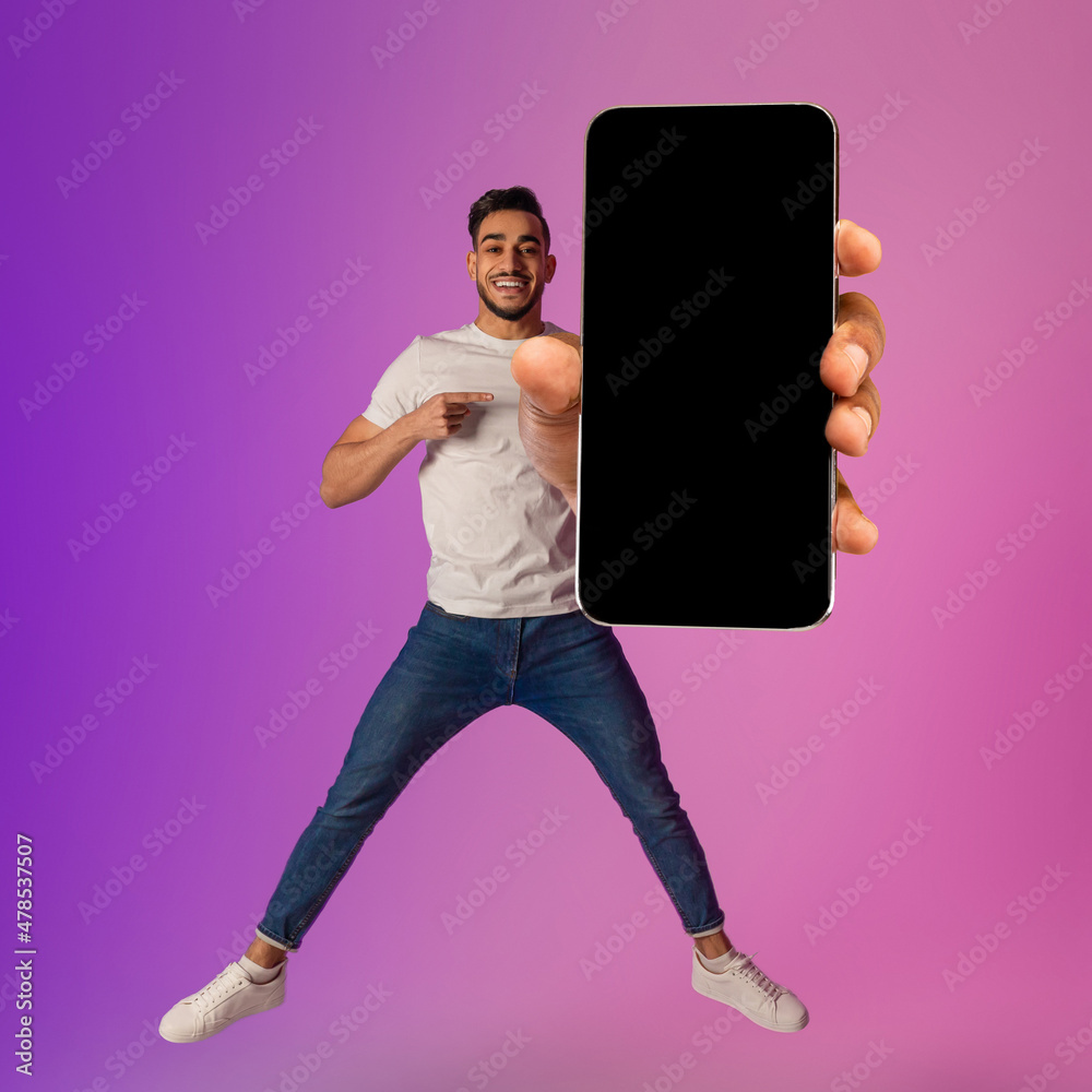 Poster handsome arab guy showing big cellphone with empty black screen, jumping in air, offering space for 