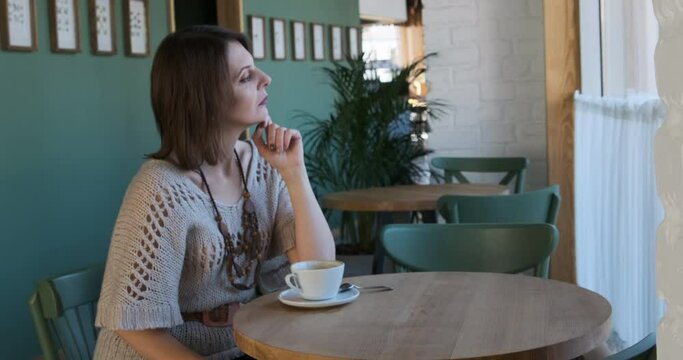 Middle-aged woman sits at table in cafe with cup of coffee and looks thoughtfully out the window. Girl came on date in a cafe and is waiting for guy