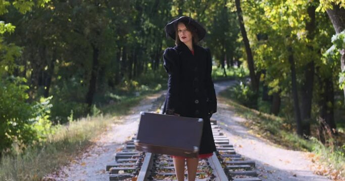 Woman dressed in the style of the 40-50s with suitcase in her hands stands on the railroad tracks. Woman in retro coat and hat