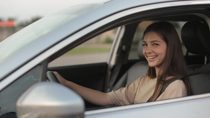 A young woman is sitting in the car and smiling. She is turning her head and looking at the camera. She is keeping her hands on the steering wheel. The camera is moving around. 4K