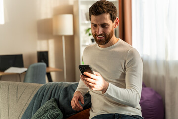 Handsome young male smiling while sitting with smartphone and looking at the screen with happy face