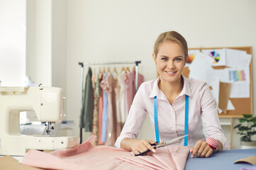 Fototapeta premium Portrait of smiling skillful young female seamstress at her workplace in sewing studio. Woman working with fabric and tailor's scissors looks at camera on background of her finished products.