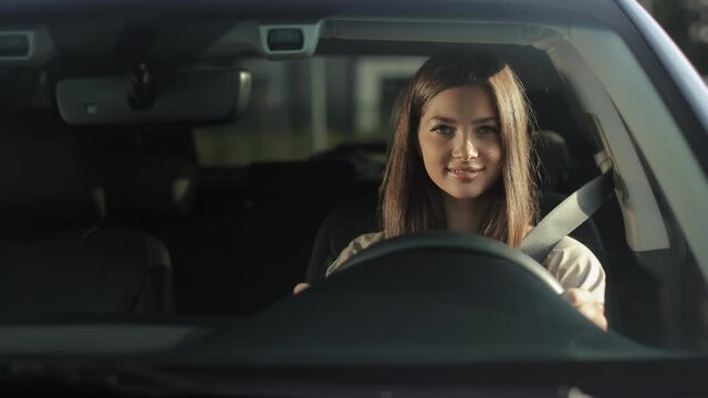 A young woman is sitting in a car in the driver's seat. She is putting on her seat belt and looking at the camera. She is smiling. 4K 50fps