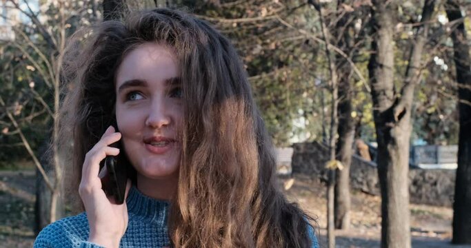 Smiling happy young girl with luxuriant brown hair talking emotionally on the phone and laughing. Sunny day in park.