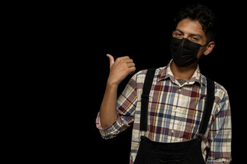 Young adult Latino male (24) with face mask points with his thumb to the right side, black background copy space.