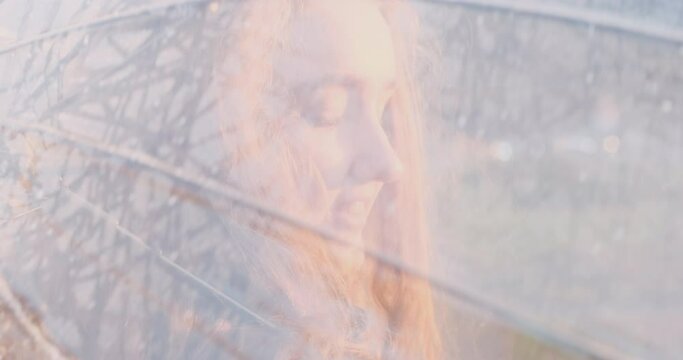 Pretty schoolgirl with chubby cheeks and long wavy hair walks around the park under an umbrella and mysteriously looks into the camera. Close up.